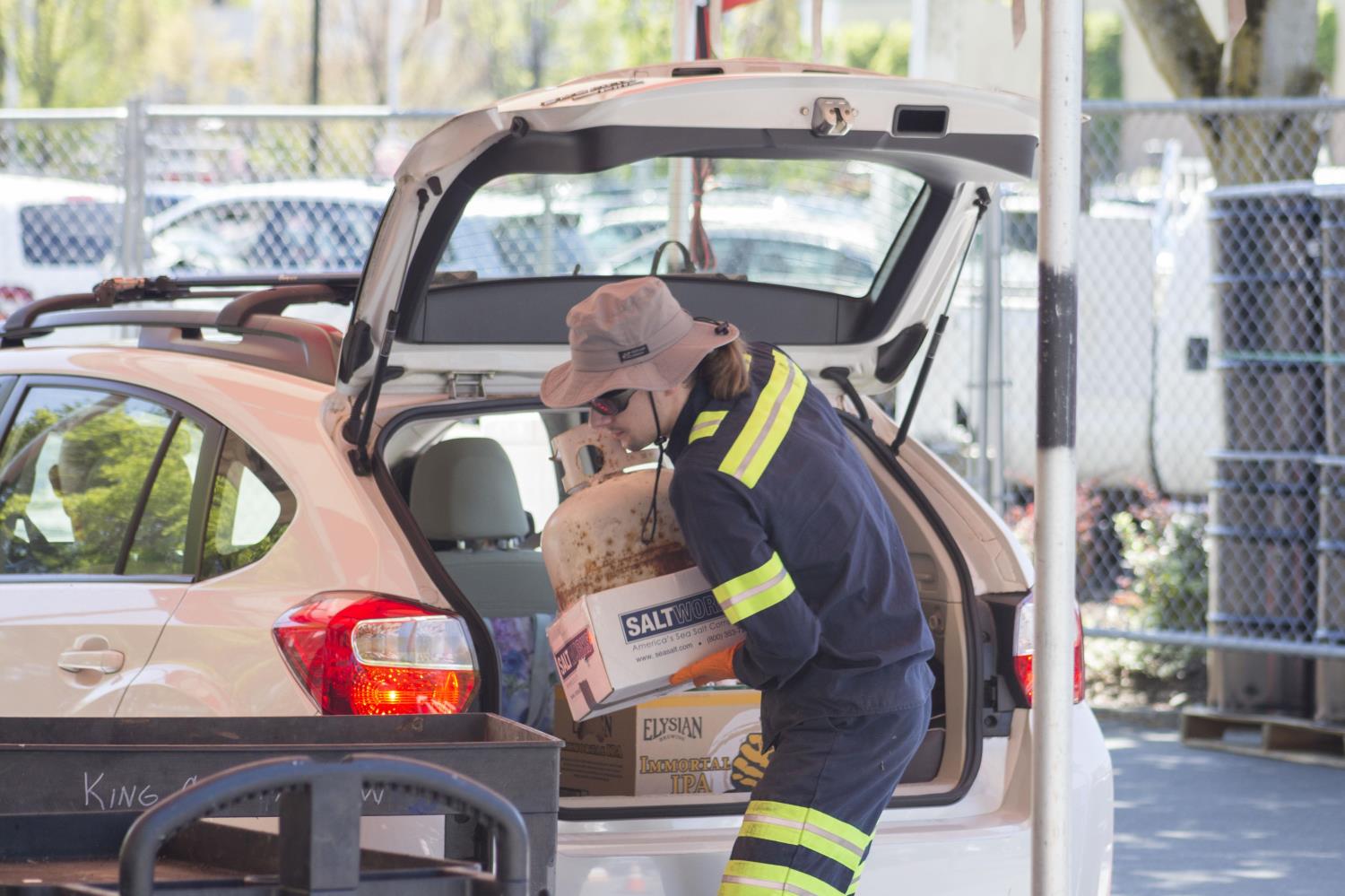 A Wastemobile staff person, wearing personal protective equipment, unloads a propane tank from the trunk of a white vehicle. 