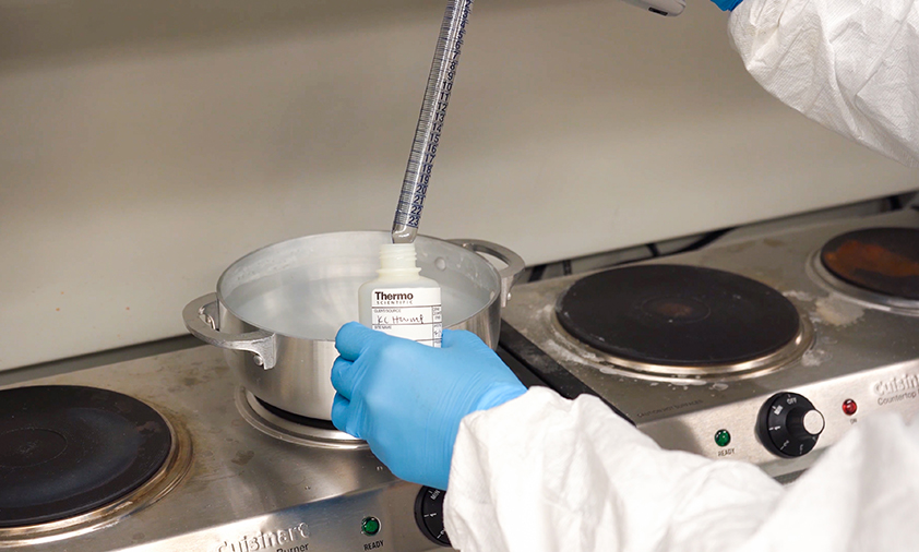 A person wearing a whitecoat and blue gloves is working in a lab. There is a pot on a burner in the background.