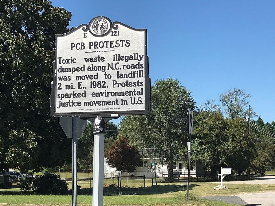 A white historic roadside sign that reads "PCB Protests: Toxic waste illegally dumped along NC roads was moved to landfill 2 mi E., 1982. Protests sparked environmental justice movement in U.S."