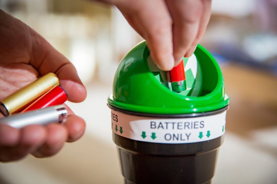 An image of a hand placing a single battery into a green topped bin labeled "Batteries Only."