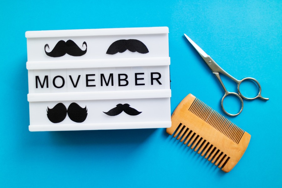 A sign that reads, "Movember" with icons of different mustache styles. Next to the sign are hair scissors and a wooden comb